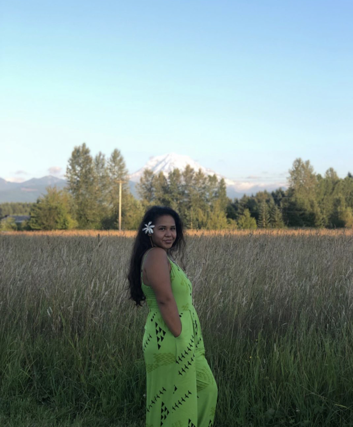 Ihilani stands in a field in a bright green dress and a flower behind her ear