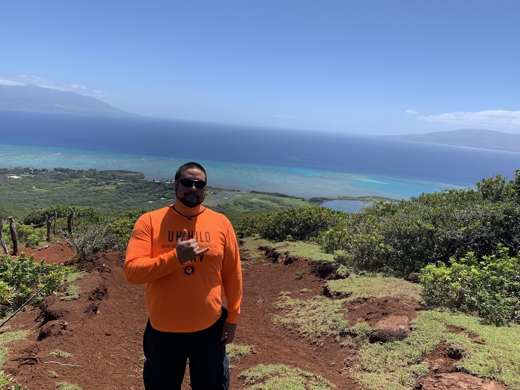Kimo throws a shaka on top of a mountain with the ocean behind him