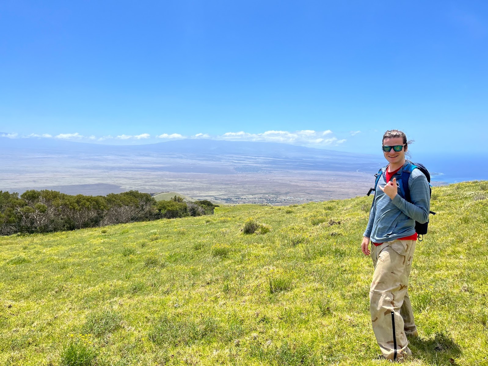 Nathaniel standing on a grassy hillside smiling and holding up a shaka with Mauna Loa and Hualālai in the background