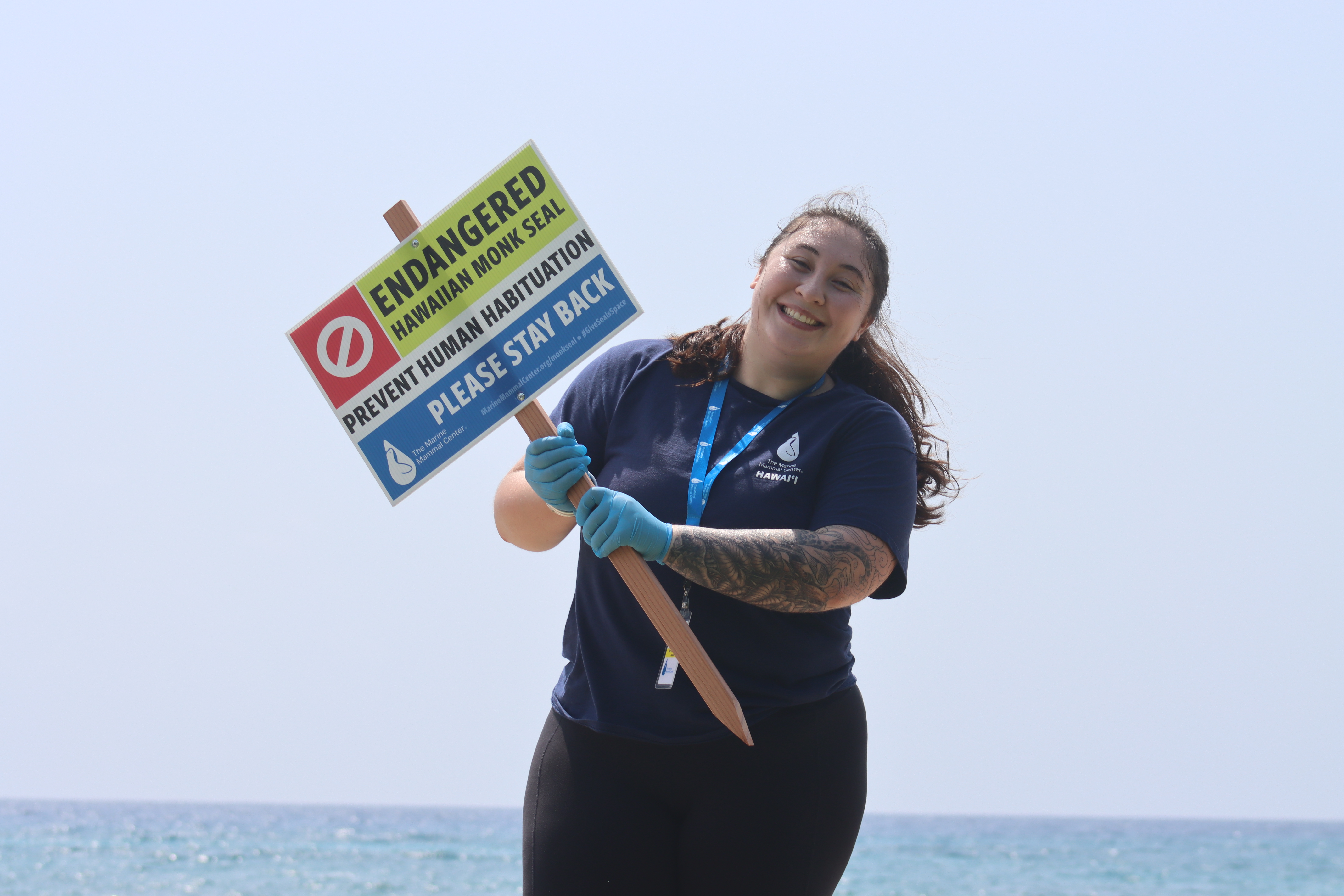 Sydney (Mina) at the beach smiling holding up a sign that says, "Endangered Hawaiian Monk Seal, Prevent Human Habituation, Please Stay Back"