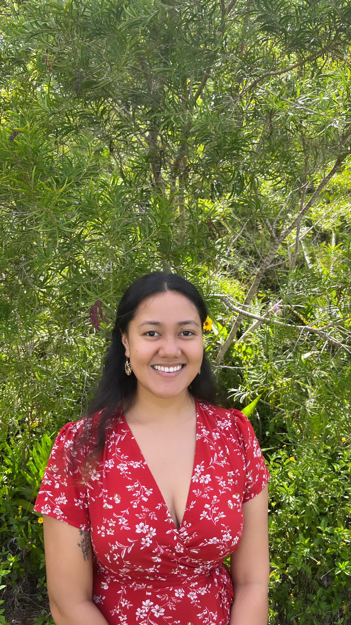 Headshot of Tuimalata smiling in a red dress and yellow flower behind her ear.