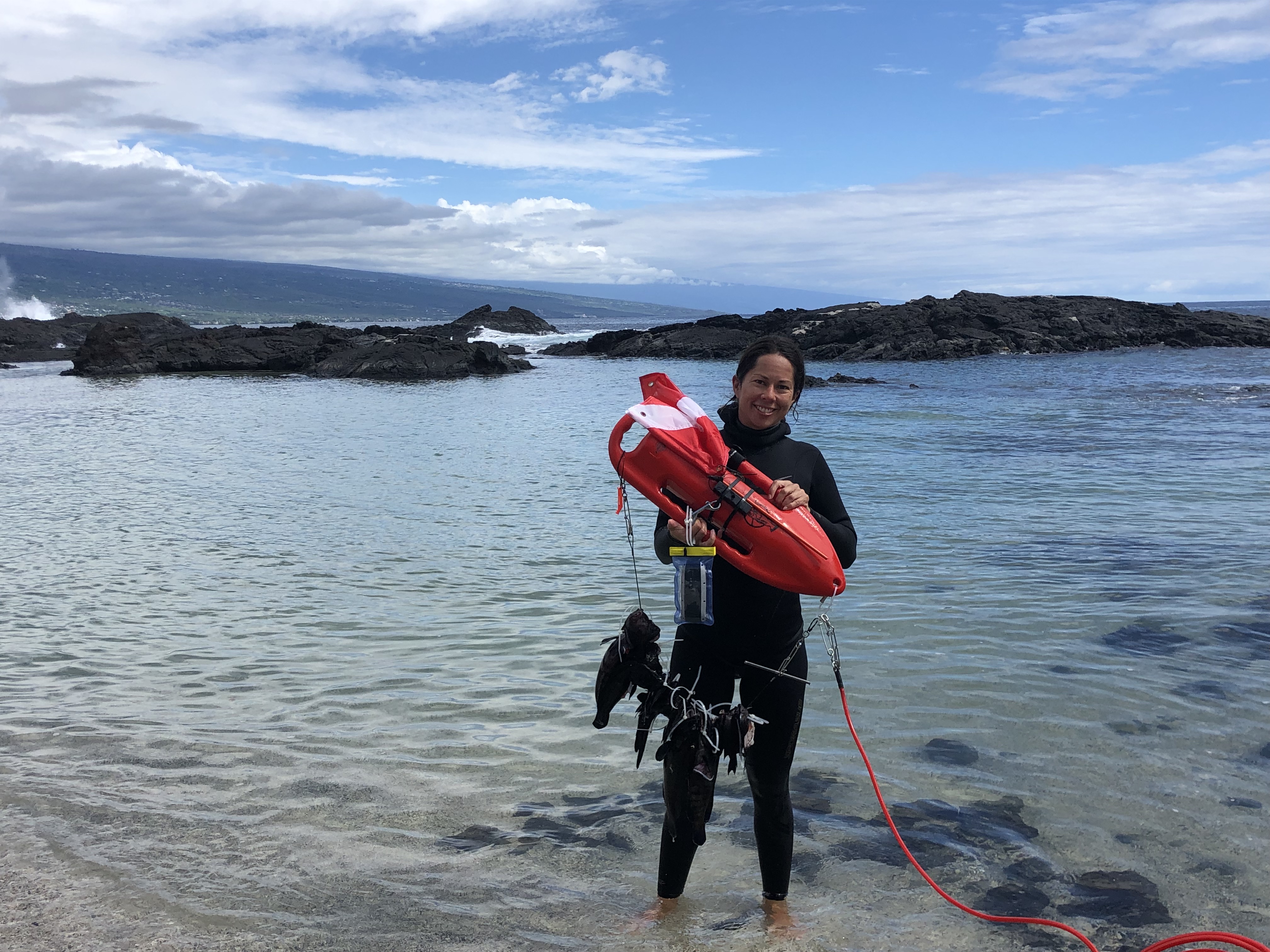 Nikola stands at the edge of the water in dive gear holding a dive buoy
