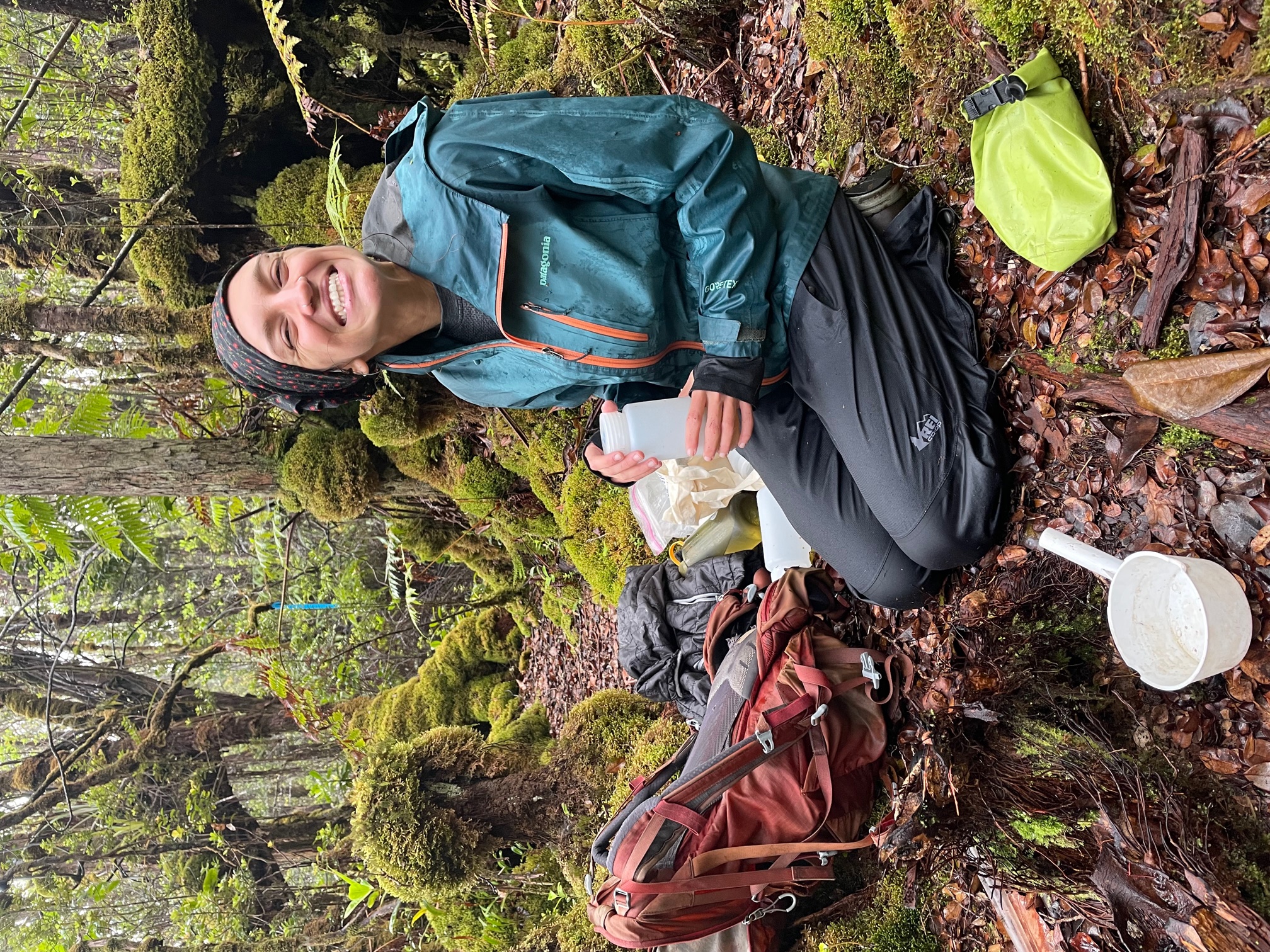 Stephanie kneels on the forest floor holding a container of liquid